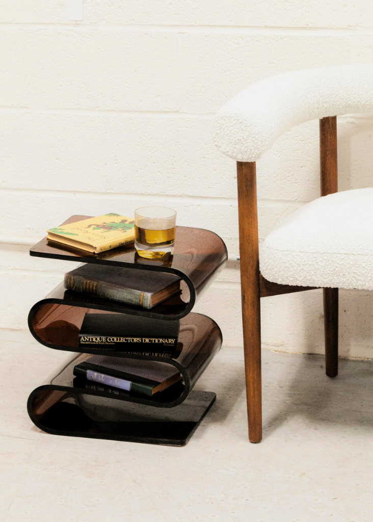 Acrylic S-Shaped Side Table with books and a glass next to a white chair.