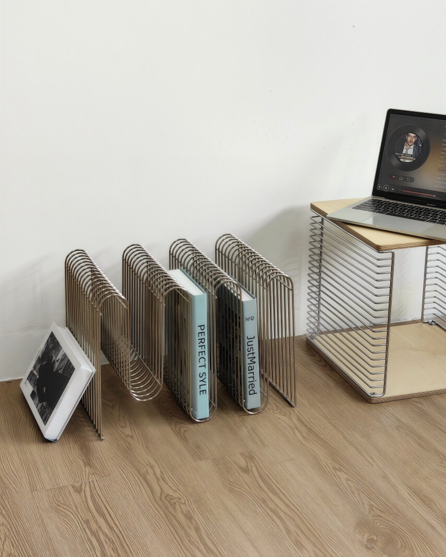 Metal curved magazine rack on a wooden floor with a laptop and tablet in the background.