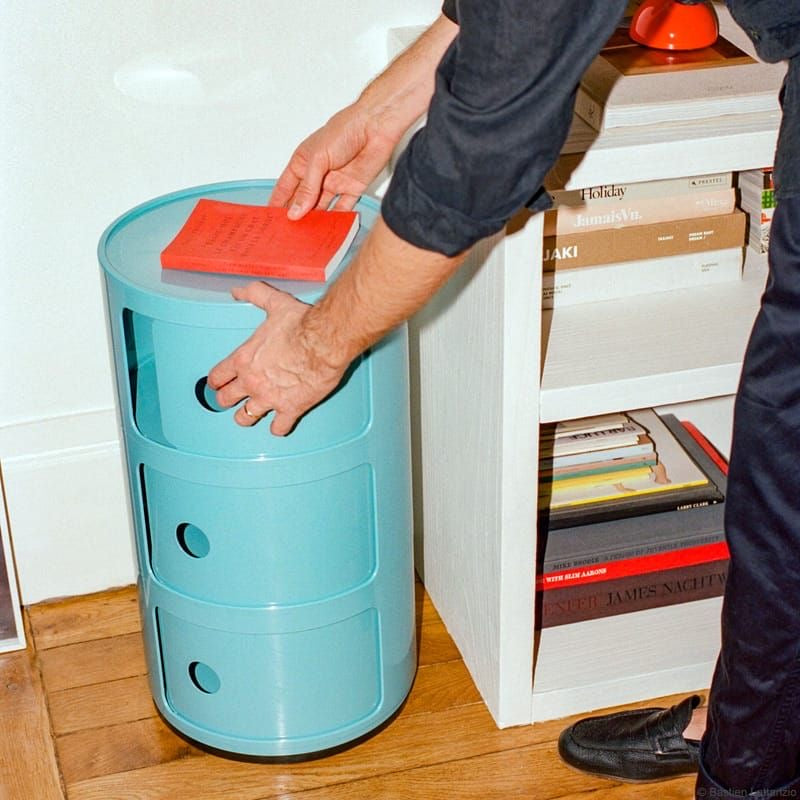 Person placing a red book on a componibili replica blue cylindrical storage unit next to a white shelf with books.