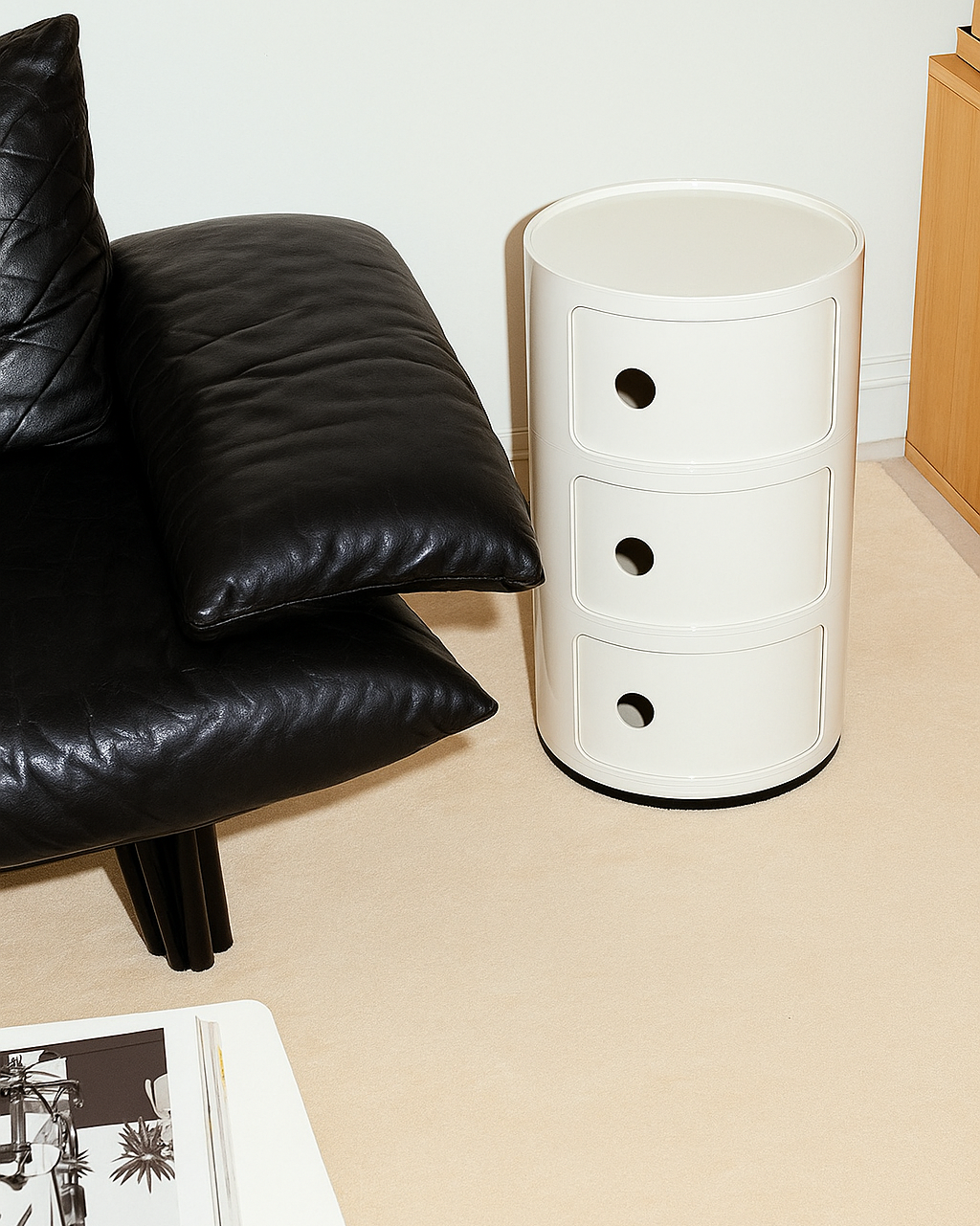 White cylindrical Componibili replica side table next to a black leather sofa on a beige floor.