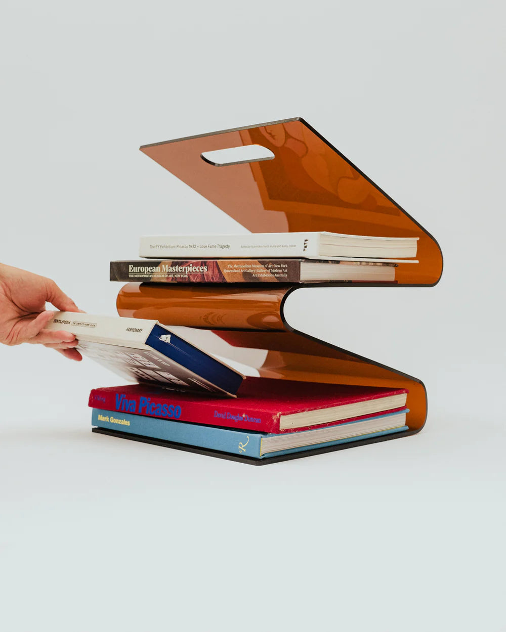 Stack of books on a transparent acrylic amber magazine rack with a hand placing another book on top.