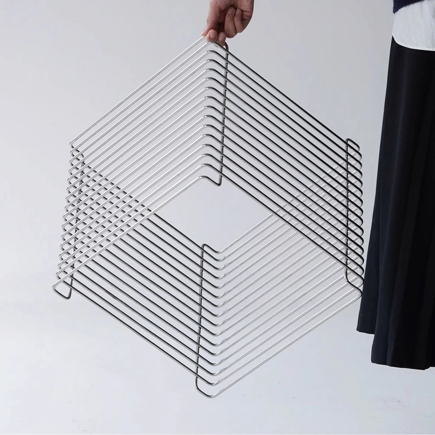 Modern wire side table made of chrome, shown on a minimalist white background. Stackable cube design with clean geometric lines. Someone in black clothes is holding the table.