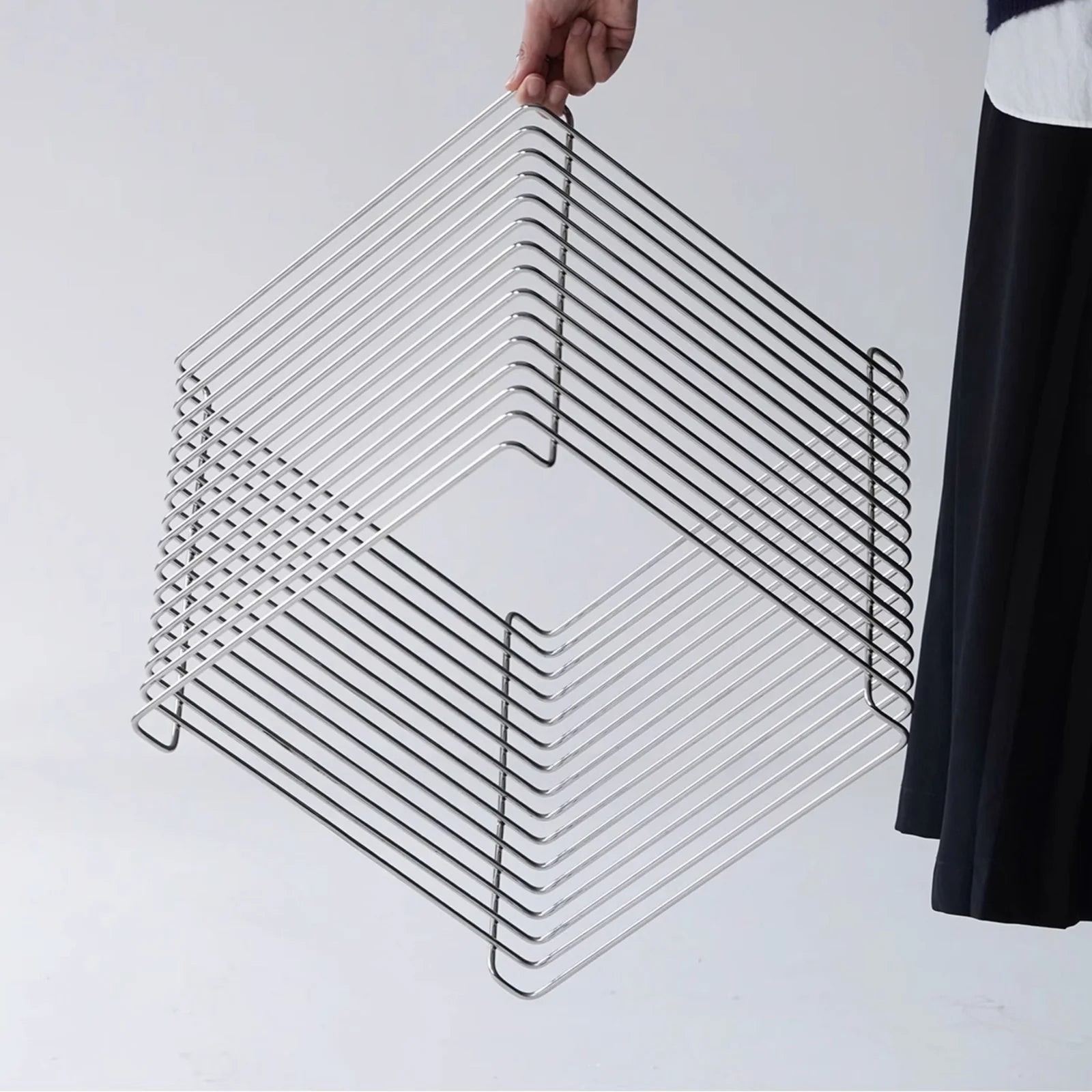 Modern wire side table made of chrome, shown on a minimalist white background. Stackable cube design with clean geometric lines. Someone in black clothes is holding the table.