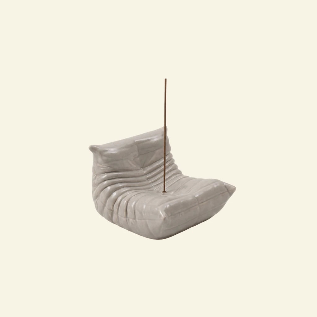 Grey Bruma incense holder shaped like a pleated lounge chair with an incense stick on a soft beige background.