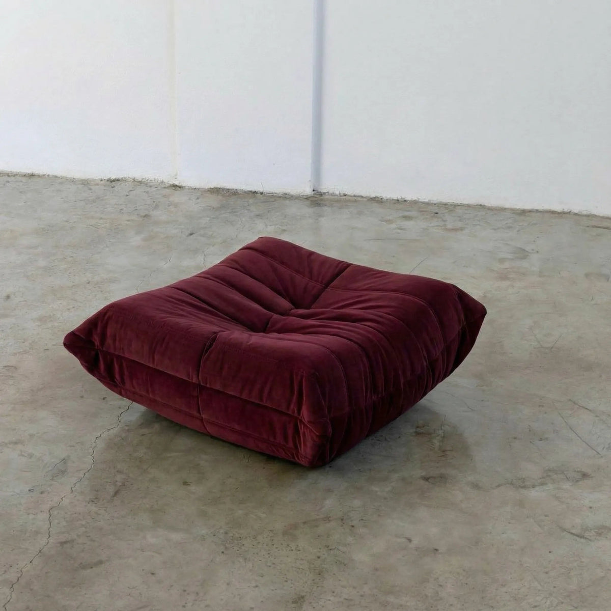Burgundy Bruma-styled ottoman in velvet suede by vibehaus on a concrete floor with white walls