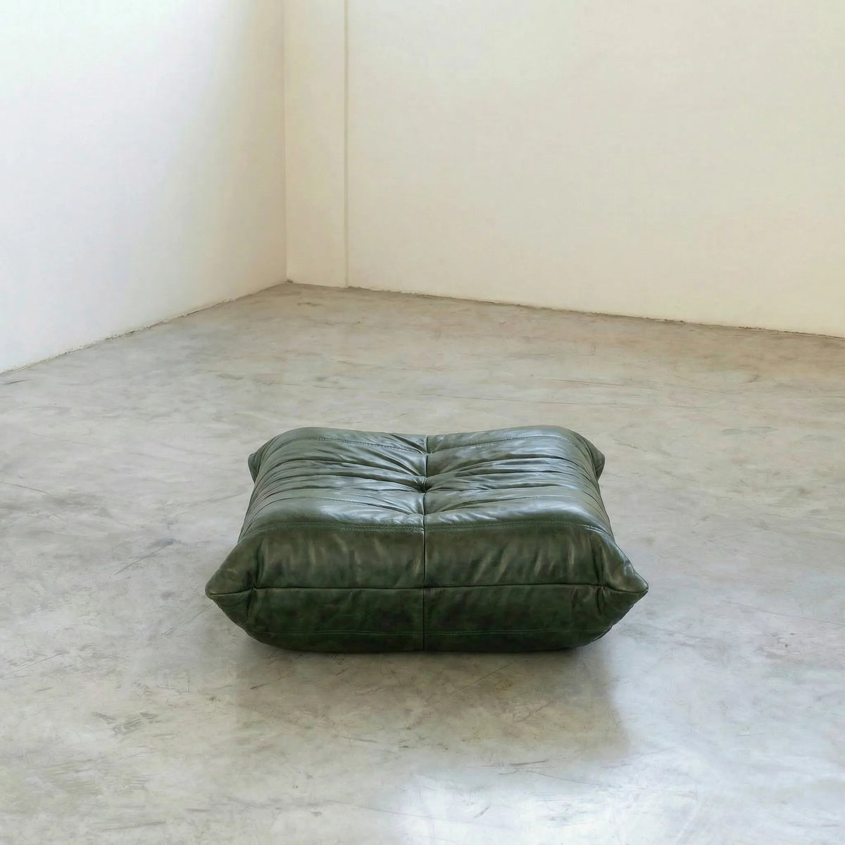 Green Bruma leather ottoman on a concrete floor with white walls.
