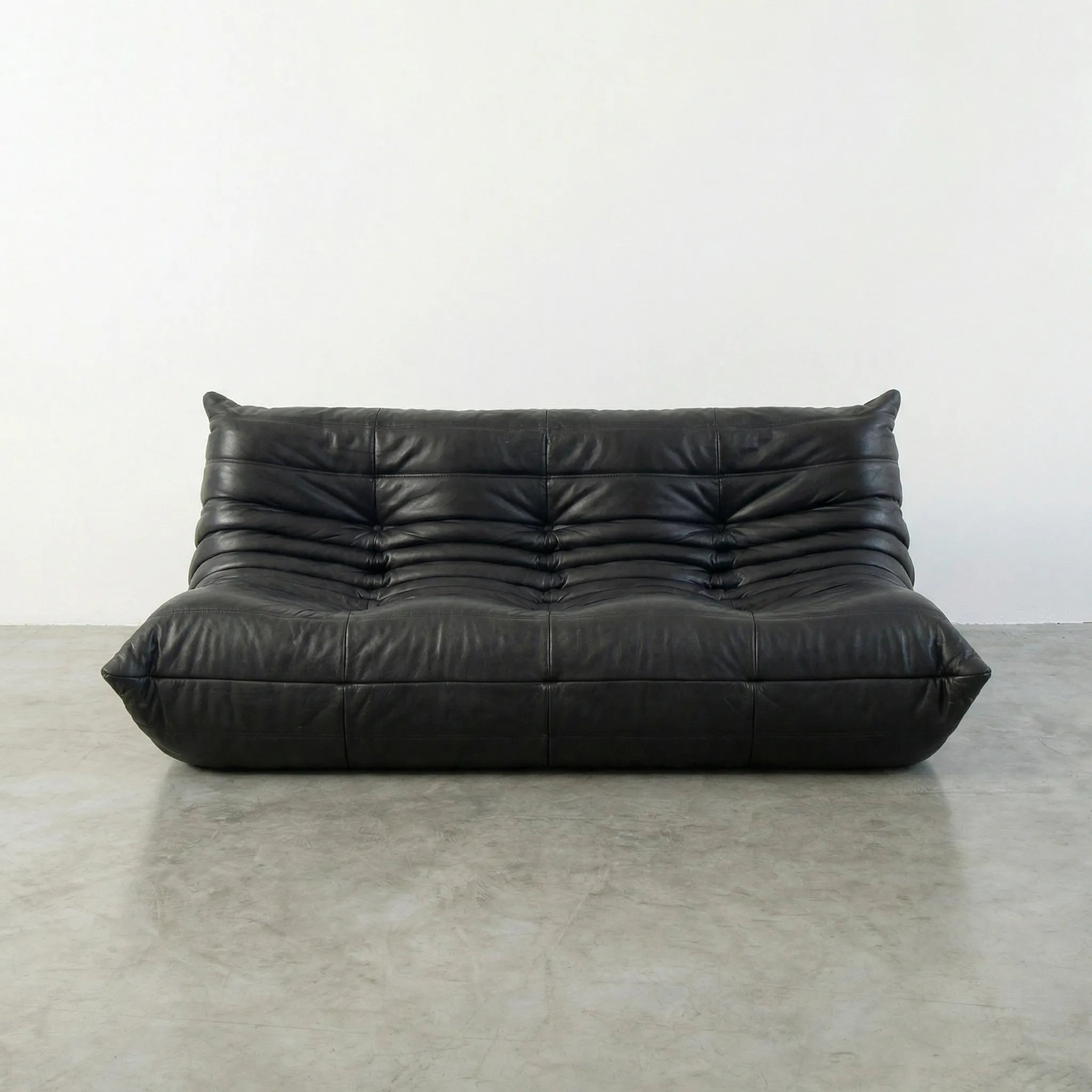 Black leather 3-seater togo sofa replica on a concrete floor with a white wall background