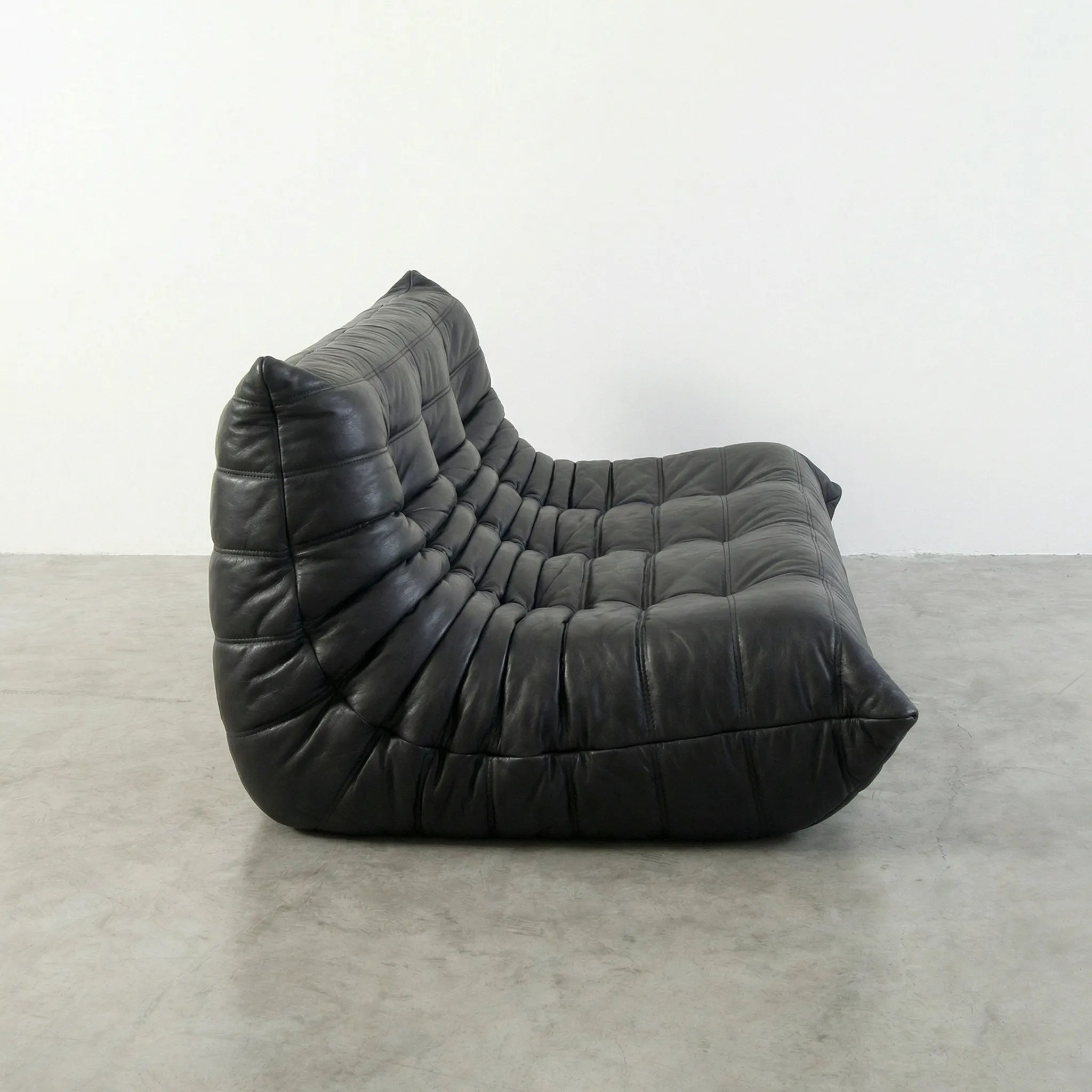 Black leather 3-seater togo sofa replica on a concrete floor with a white wall background