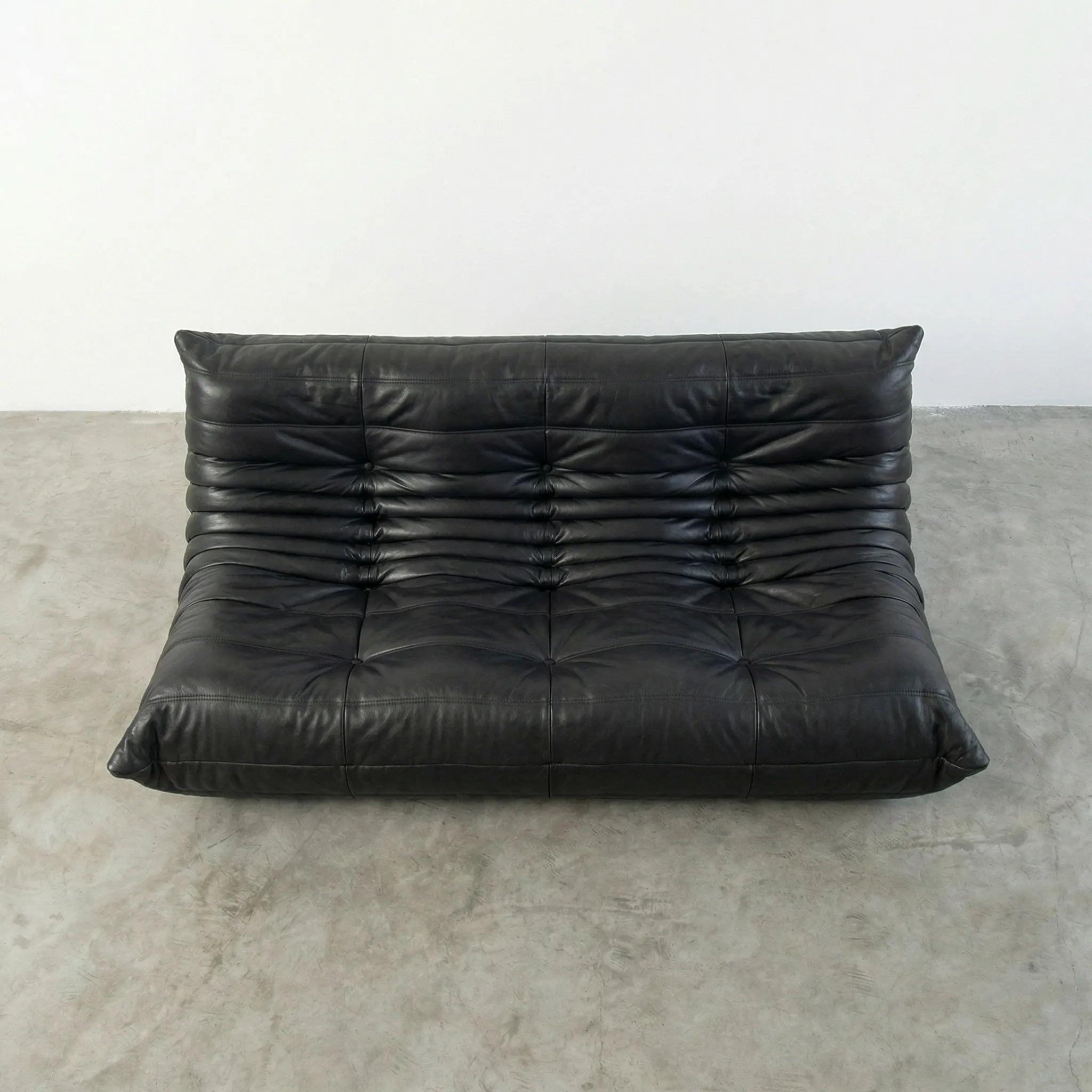 Black leather 3-seater togo sofa replica on a concrete floor with a white wall background