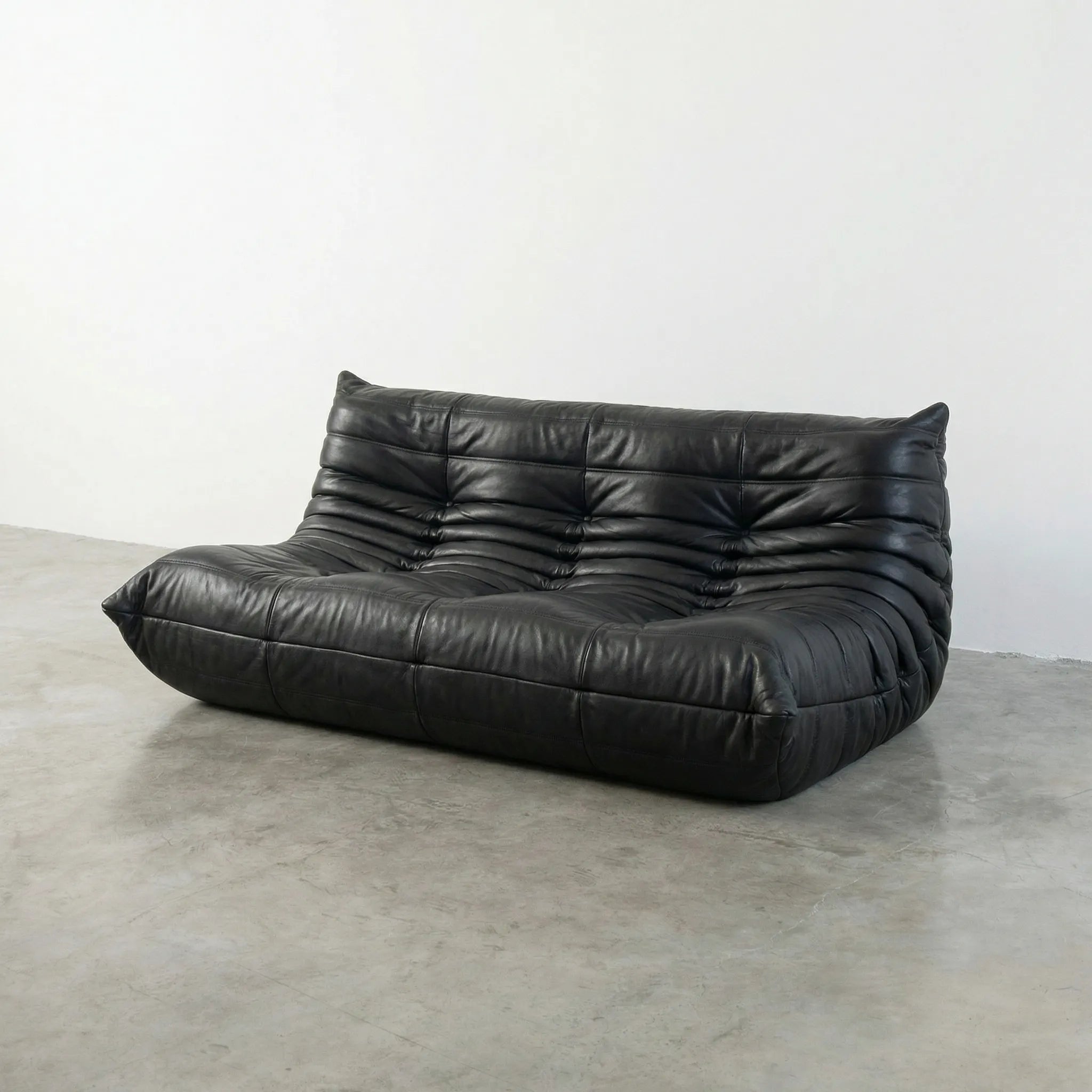 Black leather 3-seater togo sofa replica on a concrete floor with a white wall background