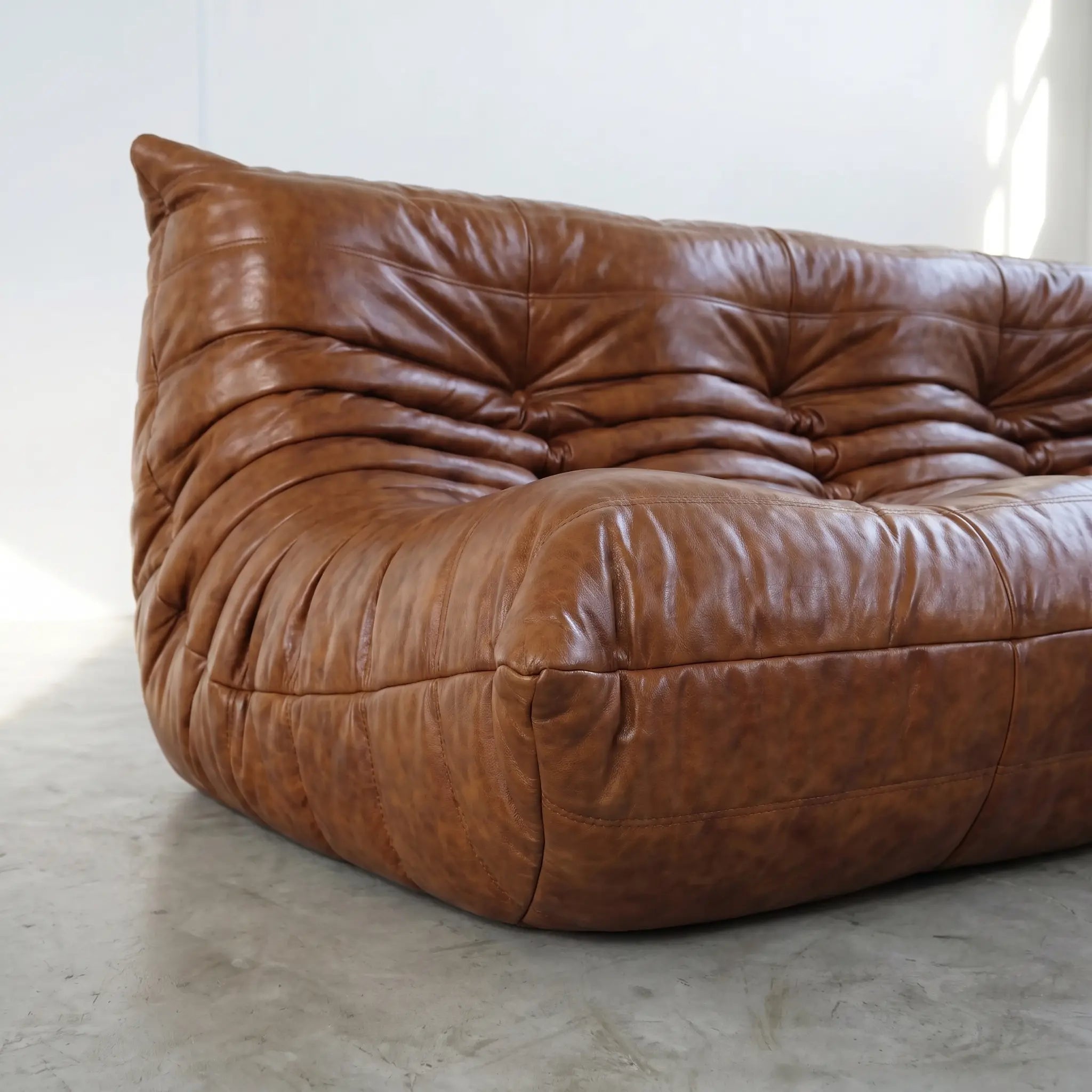A close-up of a brown leather togo sofa dupe with a heavily tufted, cloud-like silhouette, set against a minimalist white wall and concrete floor.