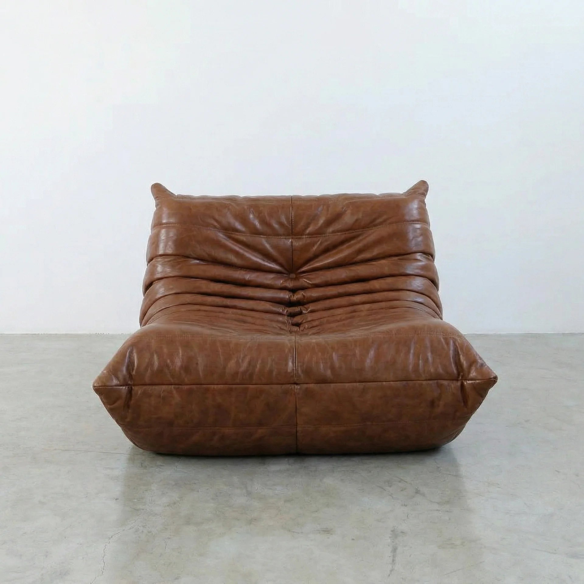 Togo Chair Dupe in brown real shiny oil wax leather on a concrete floor with a white wall background