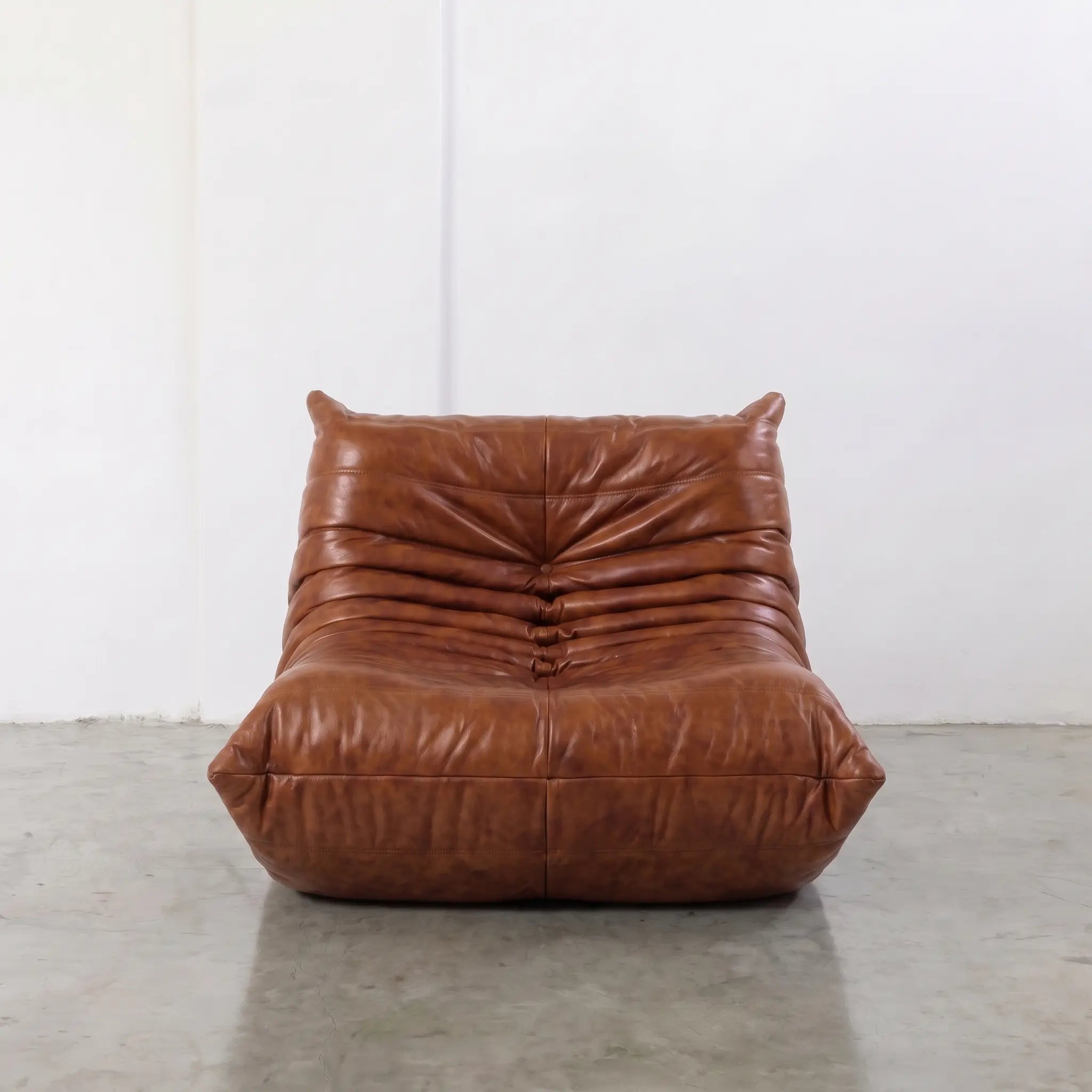 Togo Chair Dupe in brown real shiny oil wax leather on a concrete floor with a white wall background