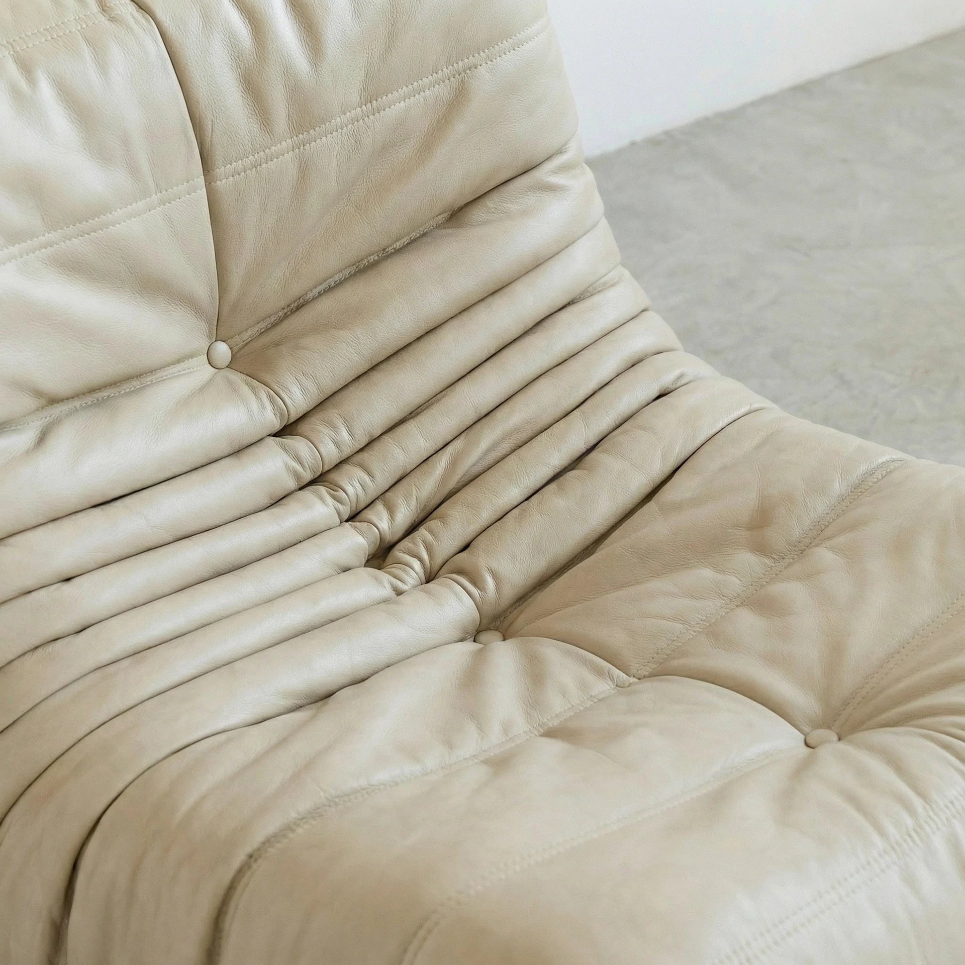close-up of the Togo Chair Dupe in cream real cowhide leather on a concrete floor with a white wall background