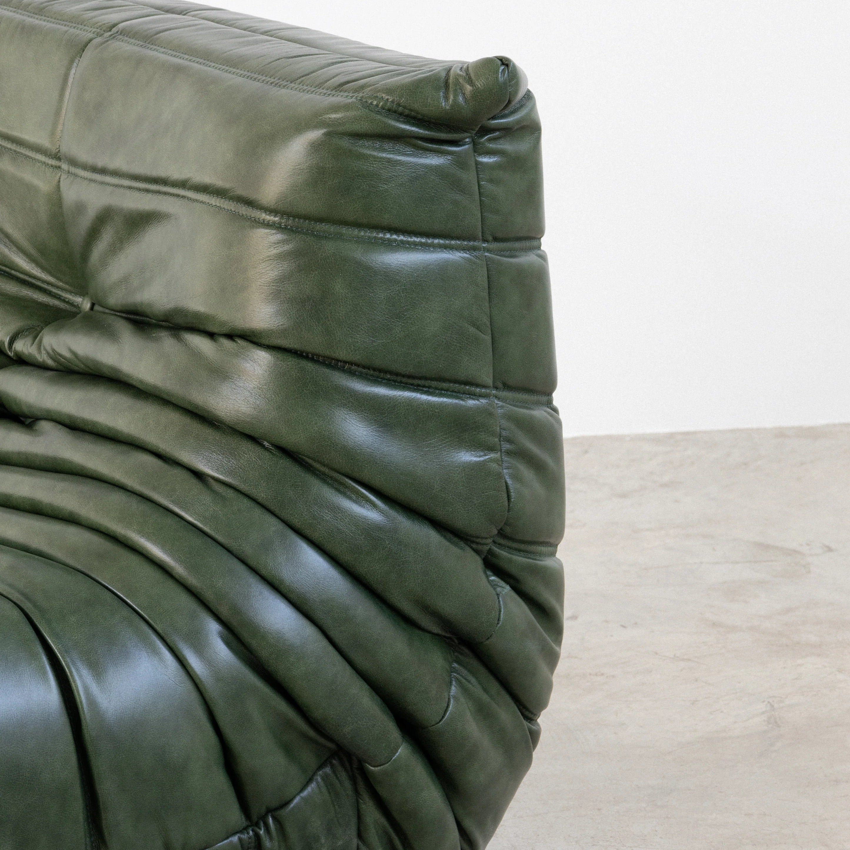 Close-up of the Togo Chair Dupe in green real shiny oil wax leather on a concrete floor with a white wall background