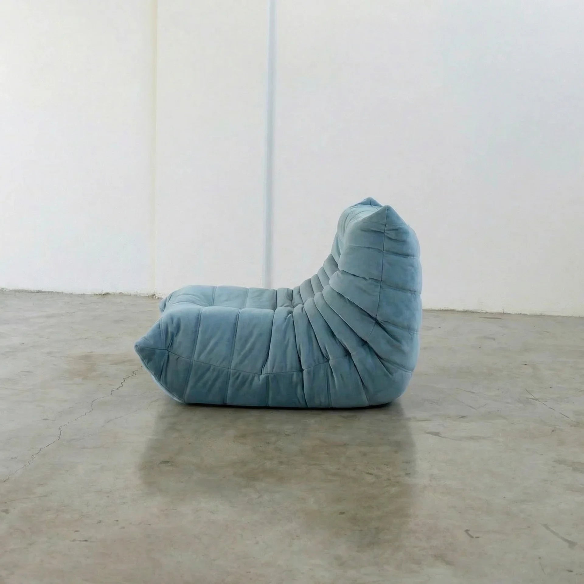 Handmade togo sofa chair as dupe in light blue velvet suede on a concrete floor with a white wall background