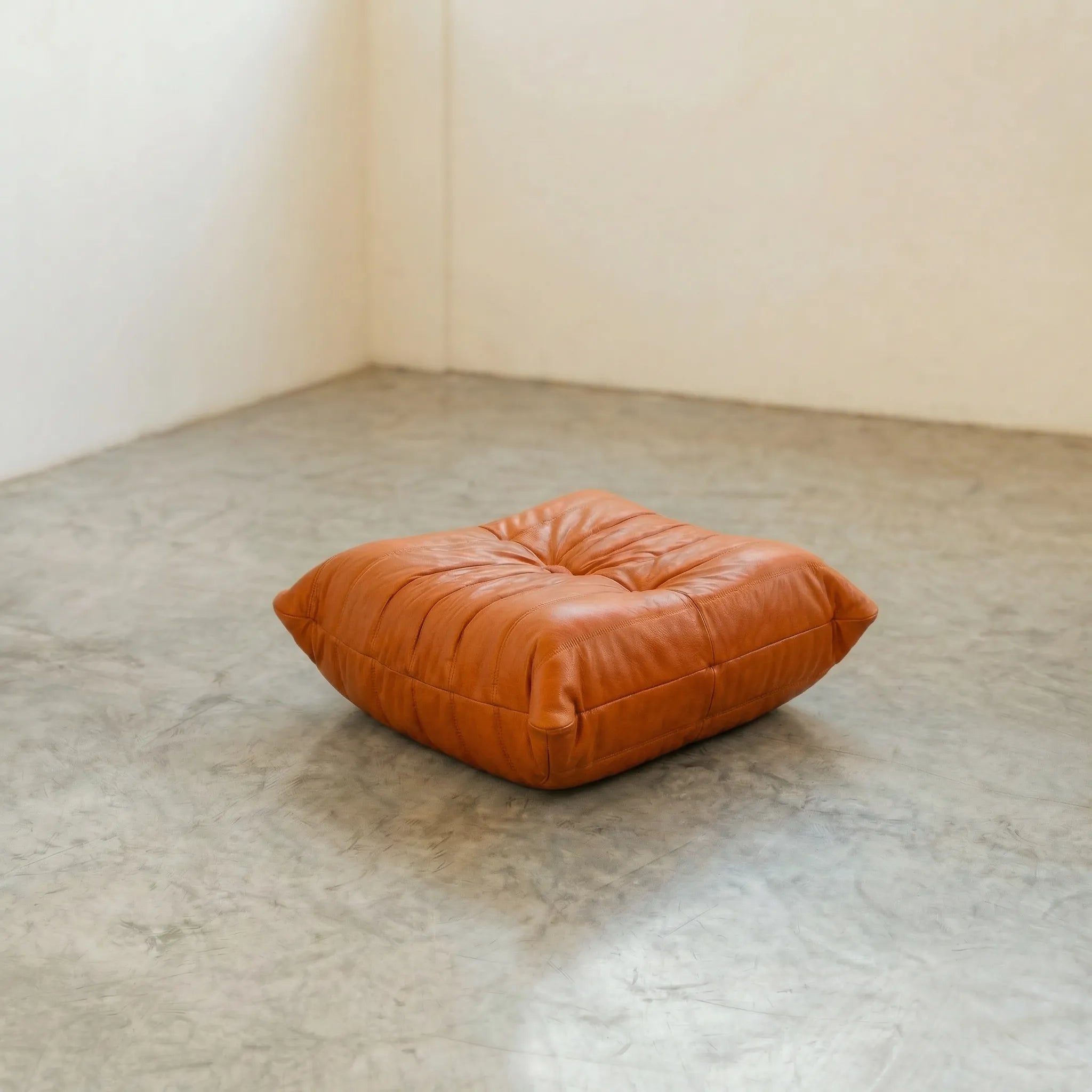 Cognac real leather togo pouf dupe on a concrete floor with a white wall background