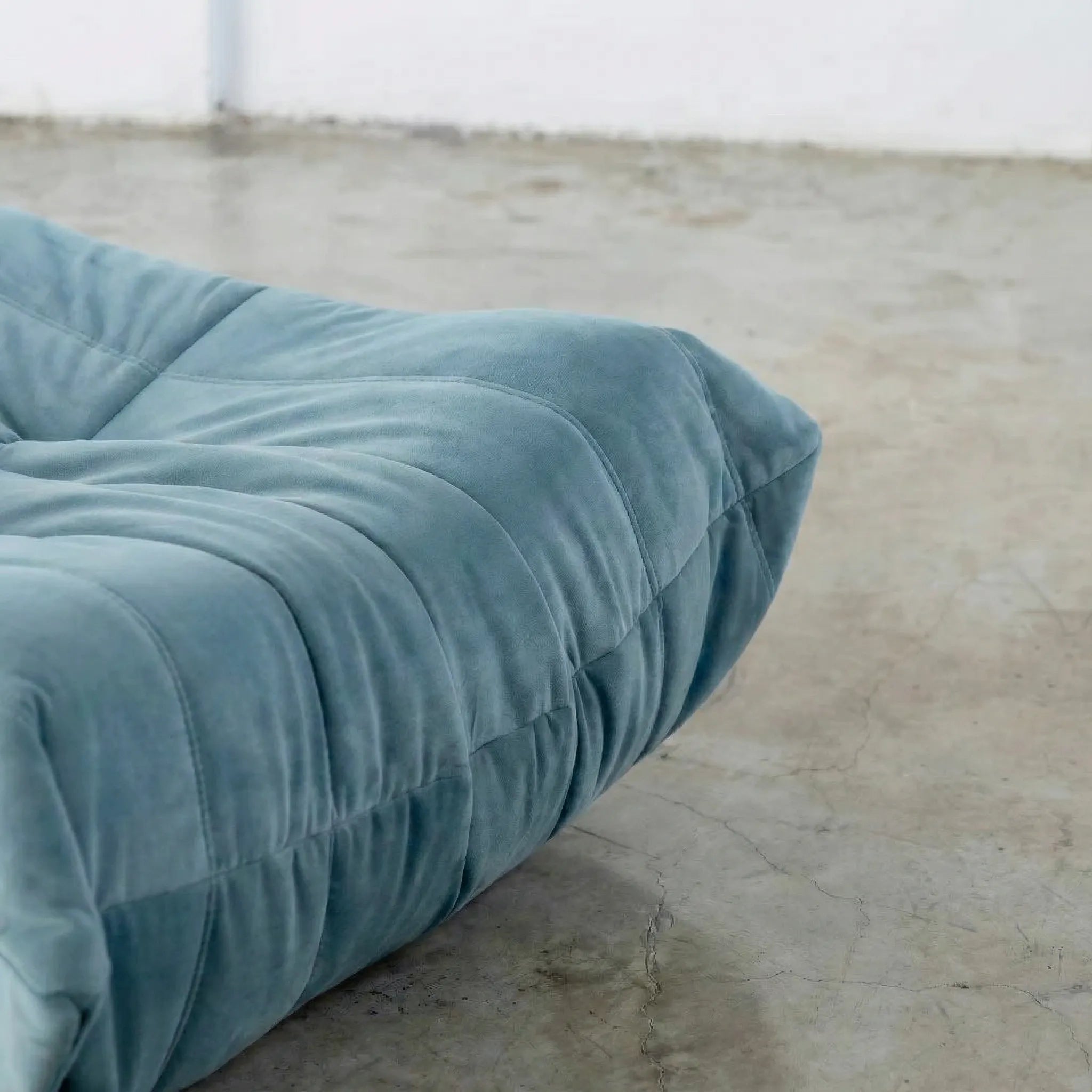 Close-up of blue togo ottoman replica in velvet suede on a concrete floor.