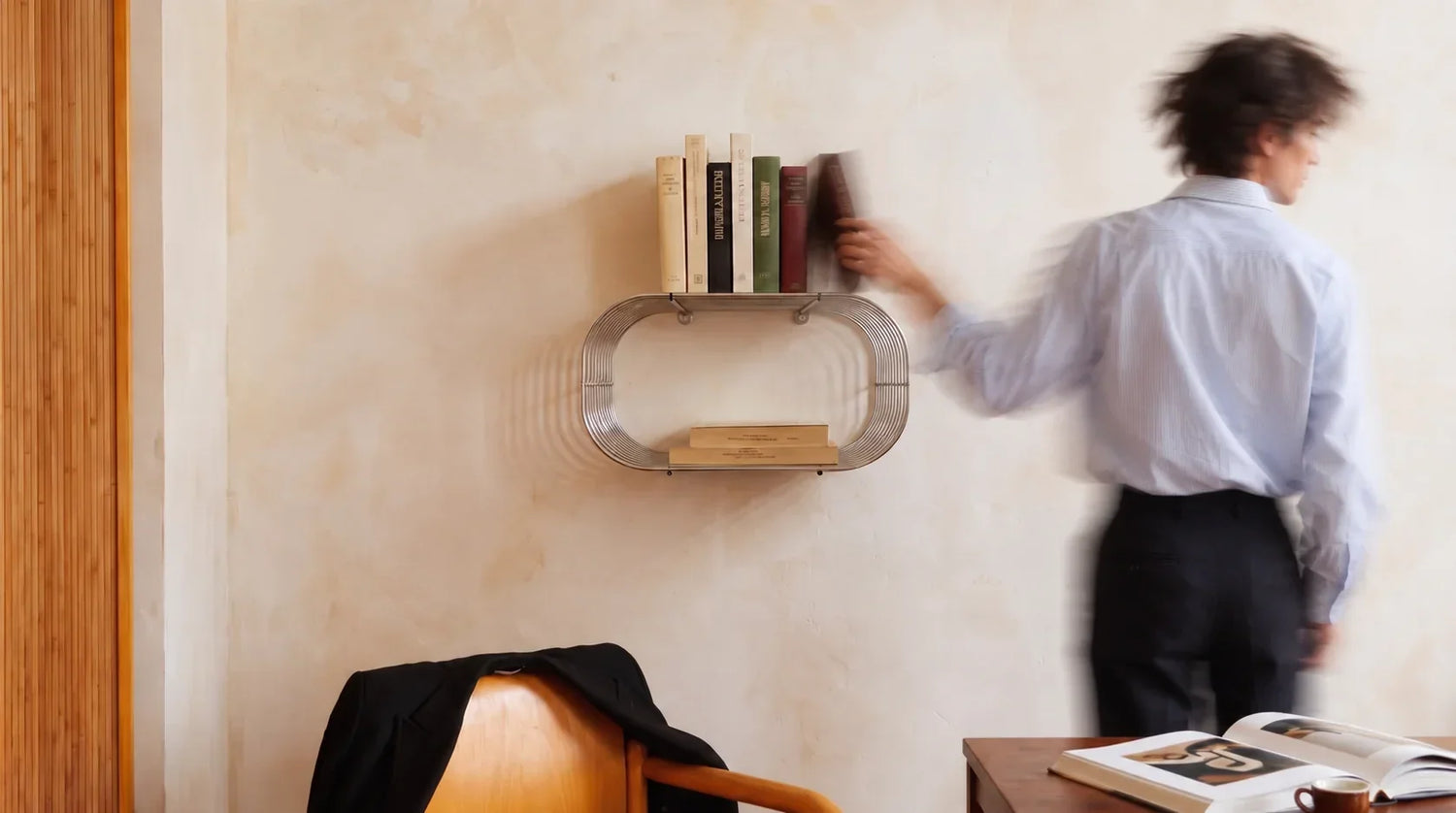 VibeHaus Chrome Wire Wall Shelf holding books on a cream wall, with a person reaching for a book.