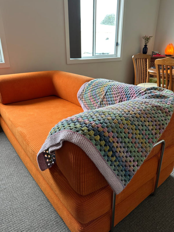Orange sofa with a multicolored knitted blanket draped over it in a room with a window and dining table.