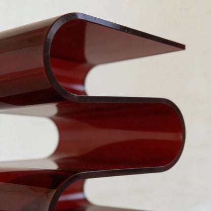 Close up of the acrylic s-shaped side table with a glossy amber red base and clear glass top on a gray background