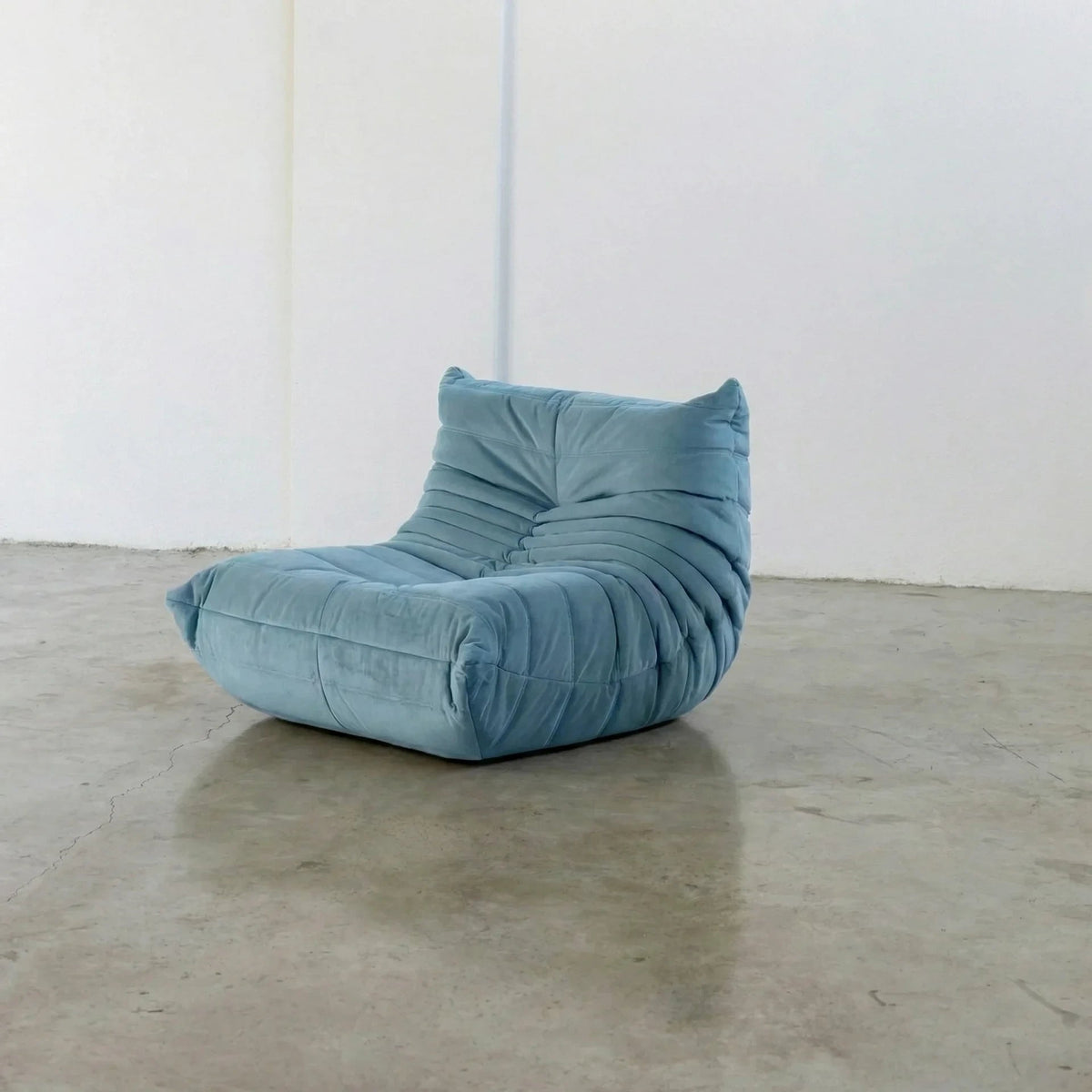 Handmade Bruma sofa chair in light blue velvet suede on a concrete floor with a white wall background