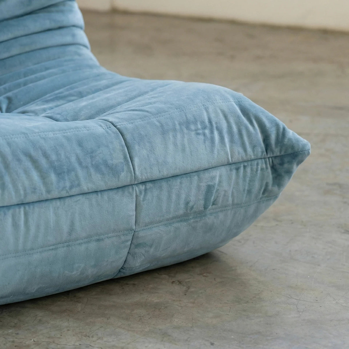 Close up of the handmade Bruma sofa chair in light blue velvet suede on a concrete floor with a white wall background