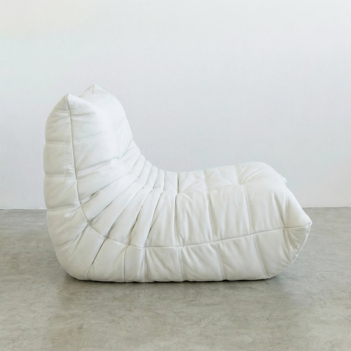 Bruma Chair in white faux leather on a concrete floor with a white wall background - shot from a side angle