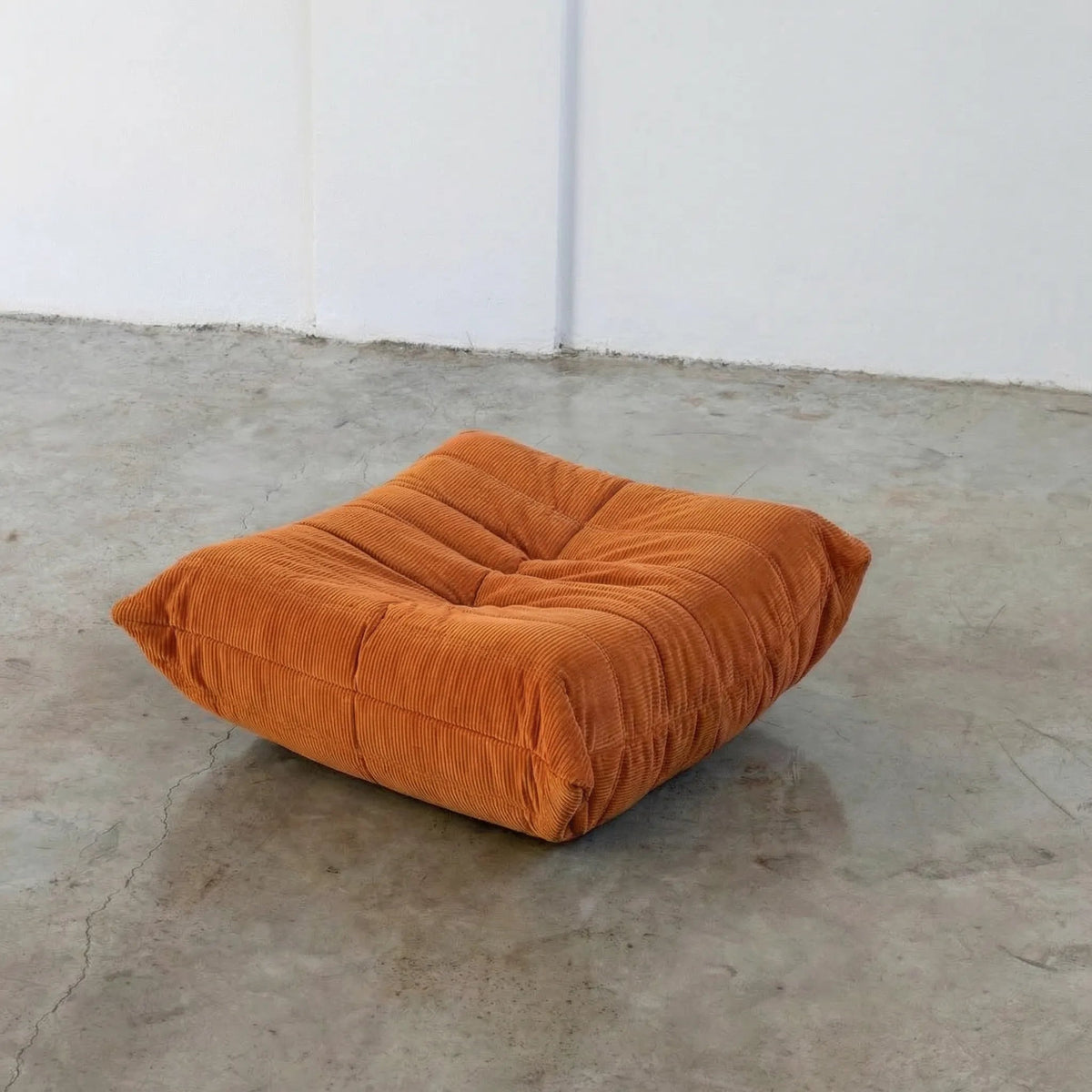 Orange Bruma-styled ottoman in corduroy by vibehaus on a concrete floor with white walls