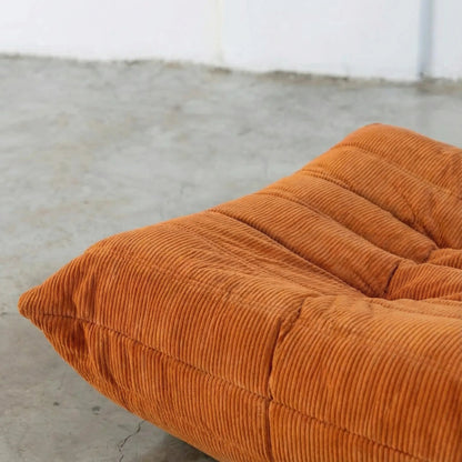 Close-up of orange Bruma ottoman in corduroy on a concrete floor.
