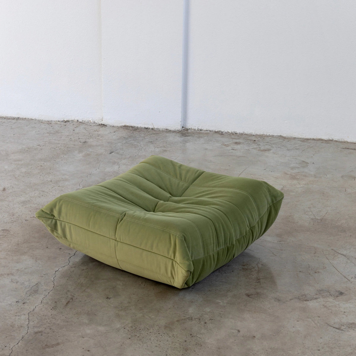 Green Bruma-styled ottoman in velvet suede by vibehaus on a concrete floor with white walls