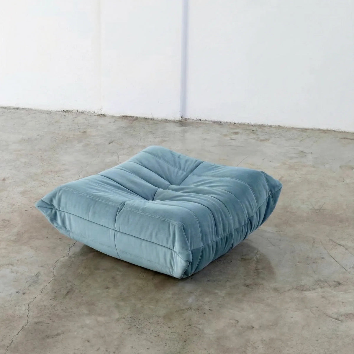 Blue Bruma-styled ottoman in velvet suede by vibehaus on a concrete floor with white walls