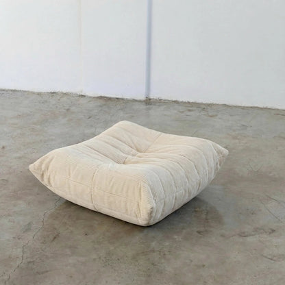 Cream Bruma-styled ottoman in corduroy by vibehaus on a concrete floor with white walls