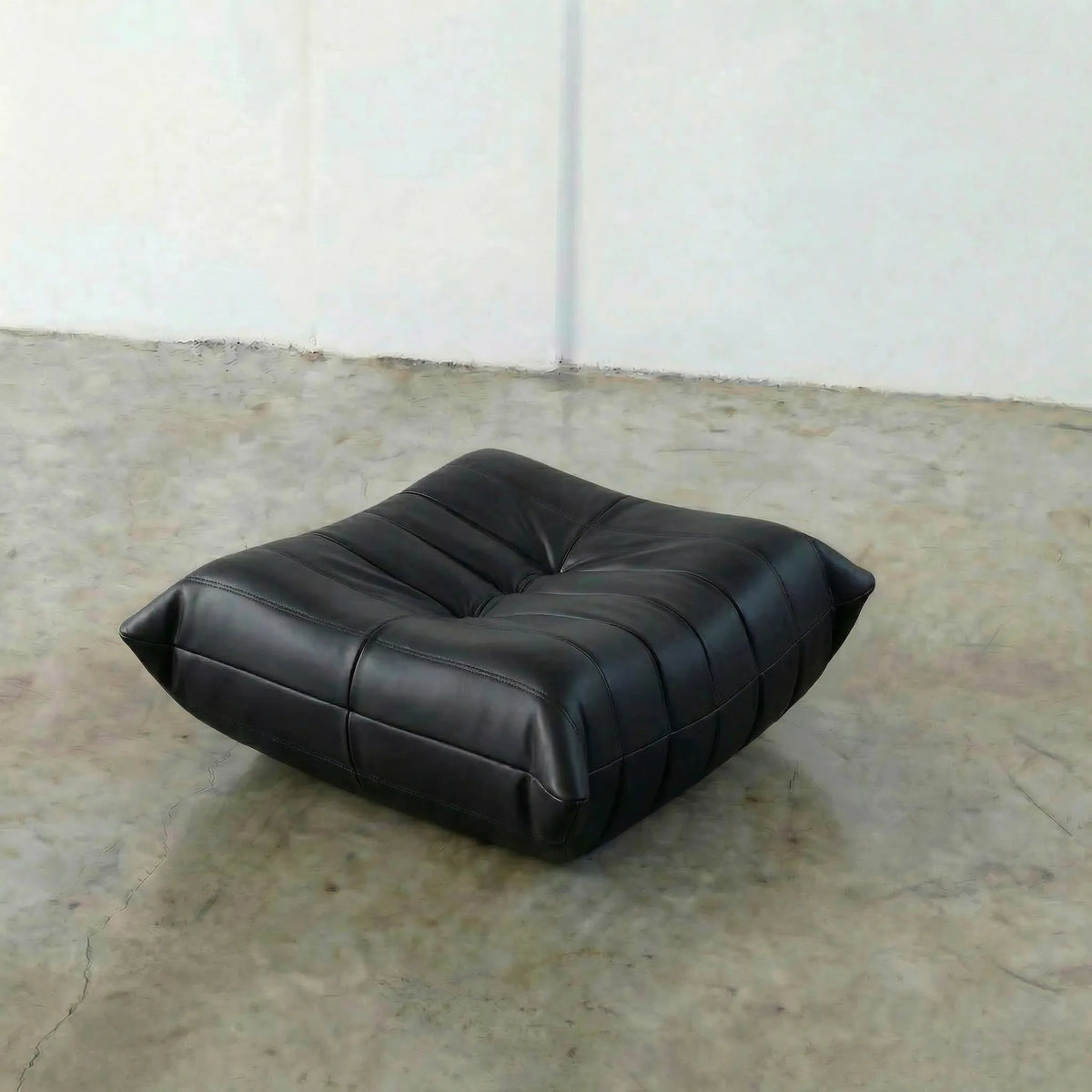 Black leather ottoman on a concrete floor with white walls.
