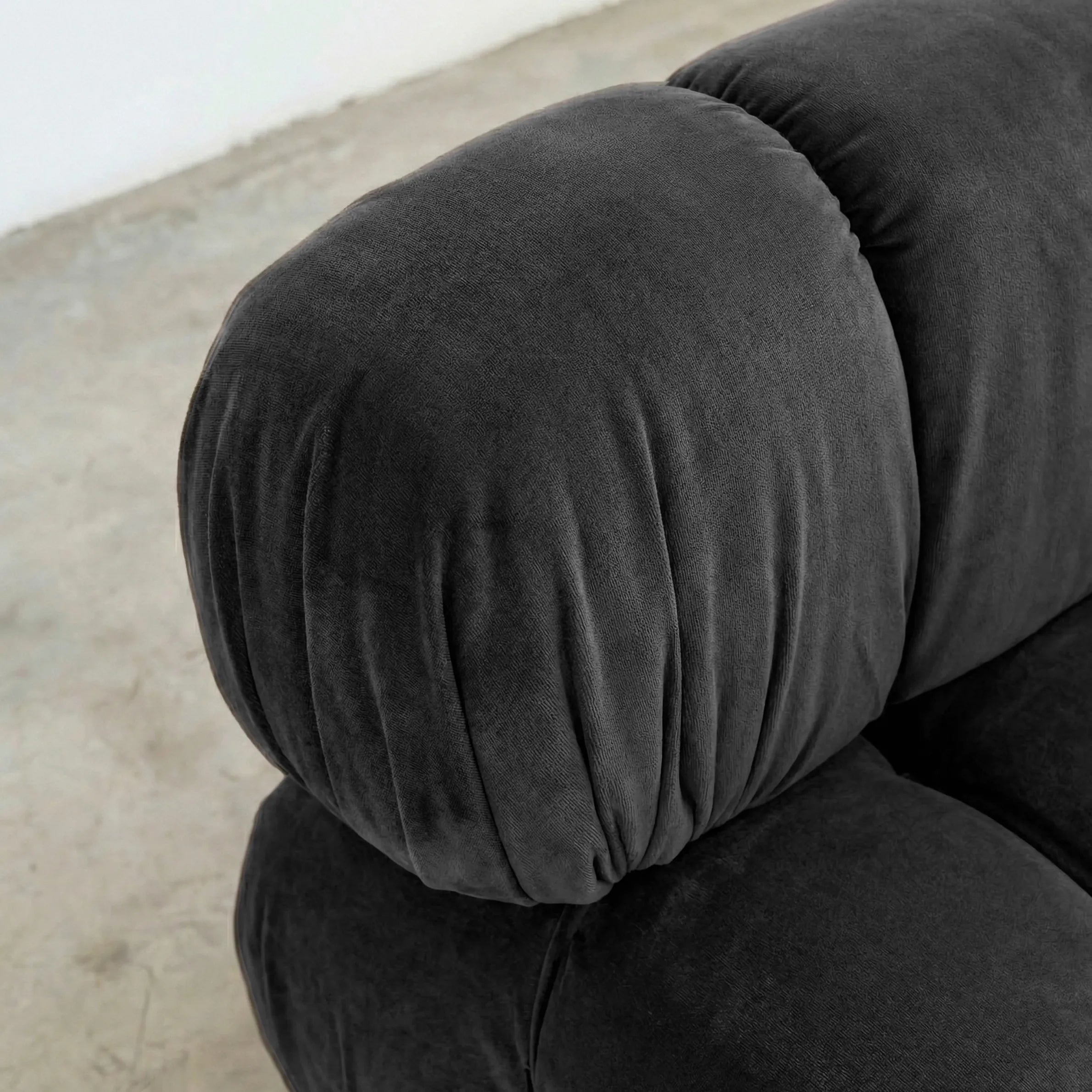 Close-up of a dark gray camaleonda dupe velvet armless section in a minimalistic room with a white wall and concrete floor.