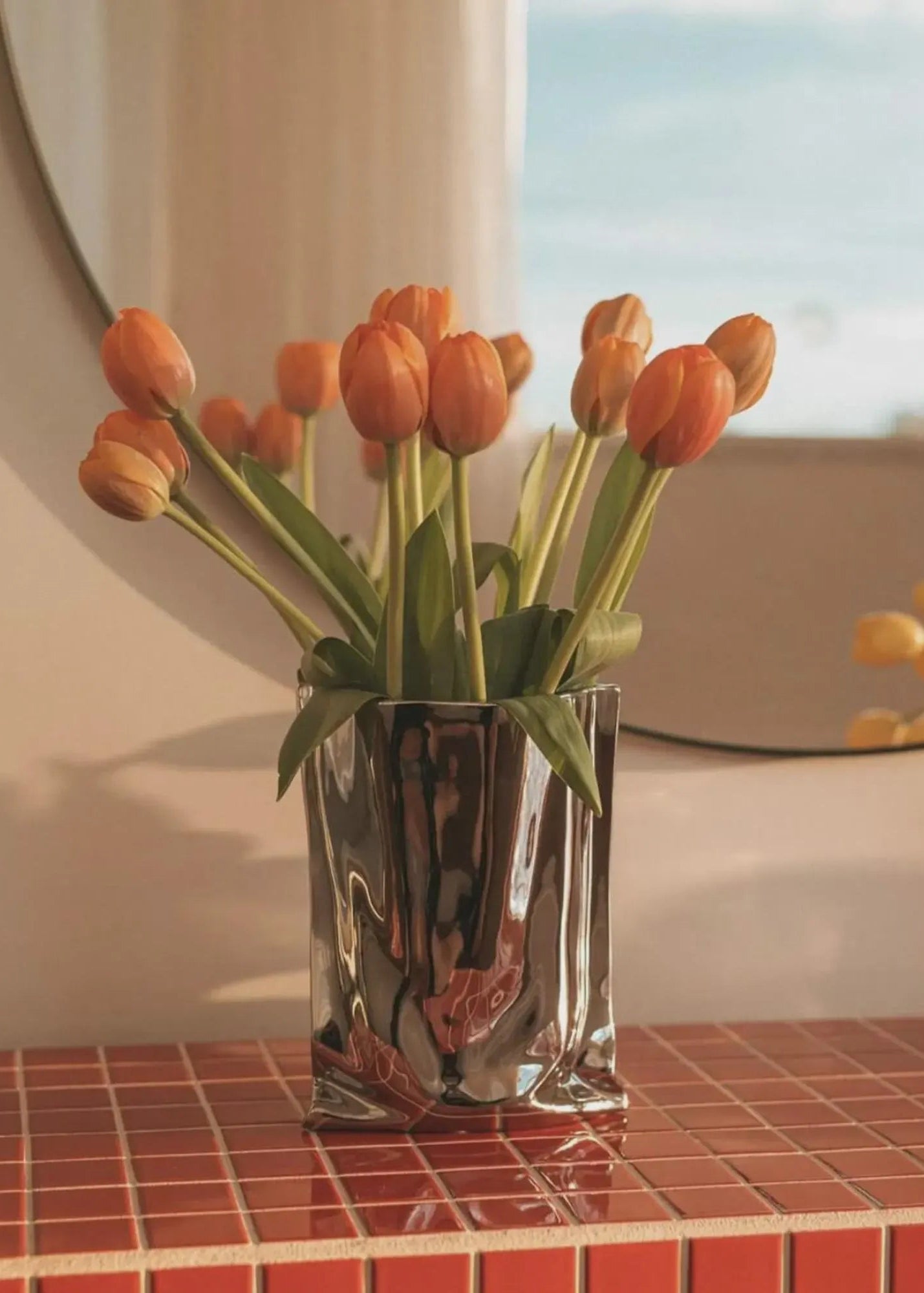 Sølv krom vase med orange tulipaner på rød flisebelagt overflade fra Vibehaus