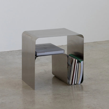 Metallic steel bauhaus side table by vibehaus with a stack of books on a plain floor.