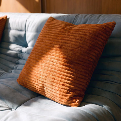 A square rust-orange corduroy throw pillow resting on a light blue tufted sofa, with warm sunlight casting shadows across its textured surface.