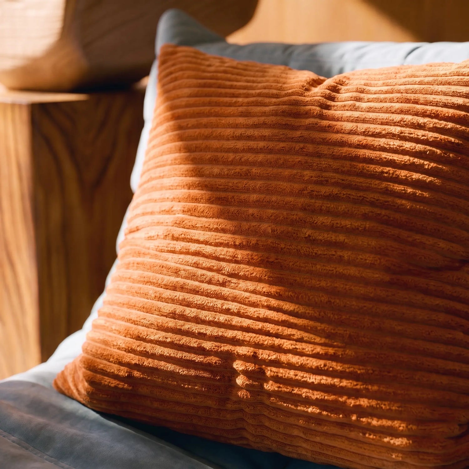 A close-up of a square orange-brown ribbed corduroy throw pillow resting on a blue-gray cushion in a warm, sunlit indoor setting.