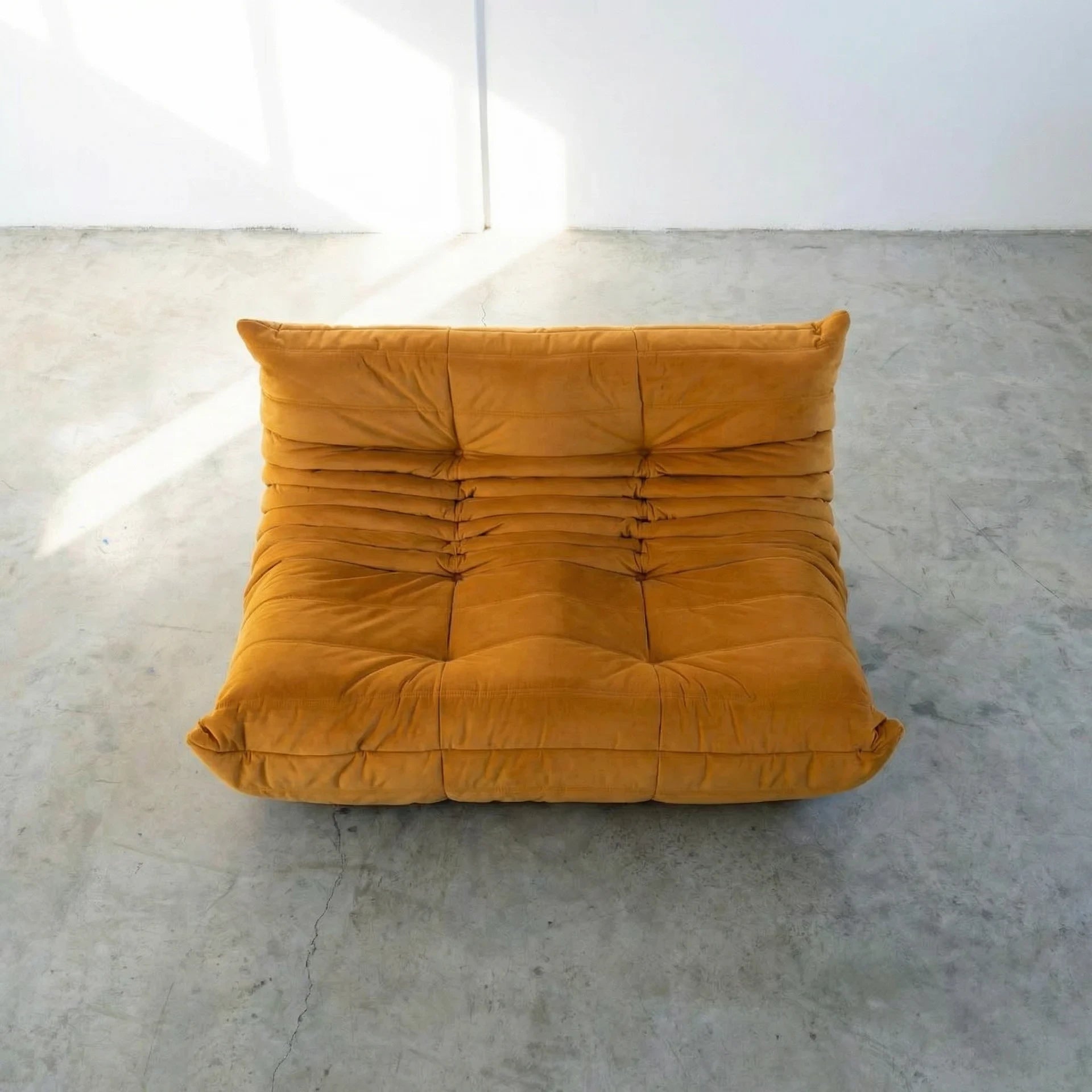 Mustard yellow dupe of the Togo 2-seater Sofa silhouette designed by michael ducaroy for ligne roset – custom-made yellow velvet seating for modern interiors on a concrete floor with white walls