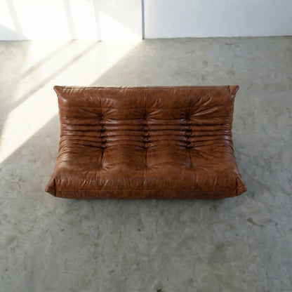 A brown togo dupe sofa in leather, photographed from above on a light concrete floor with bright sunlight casting shadows.