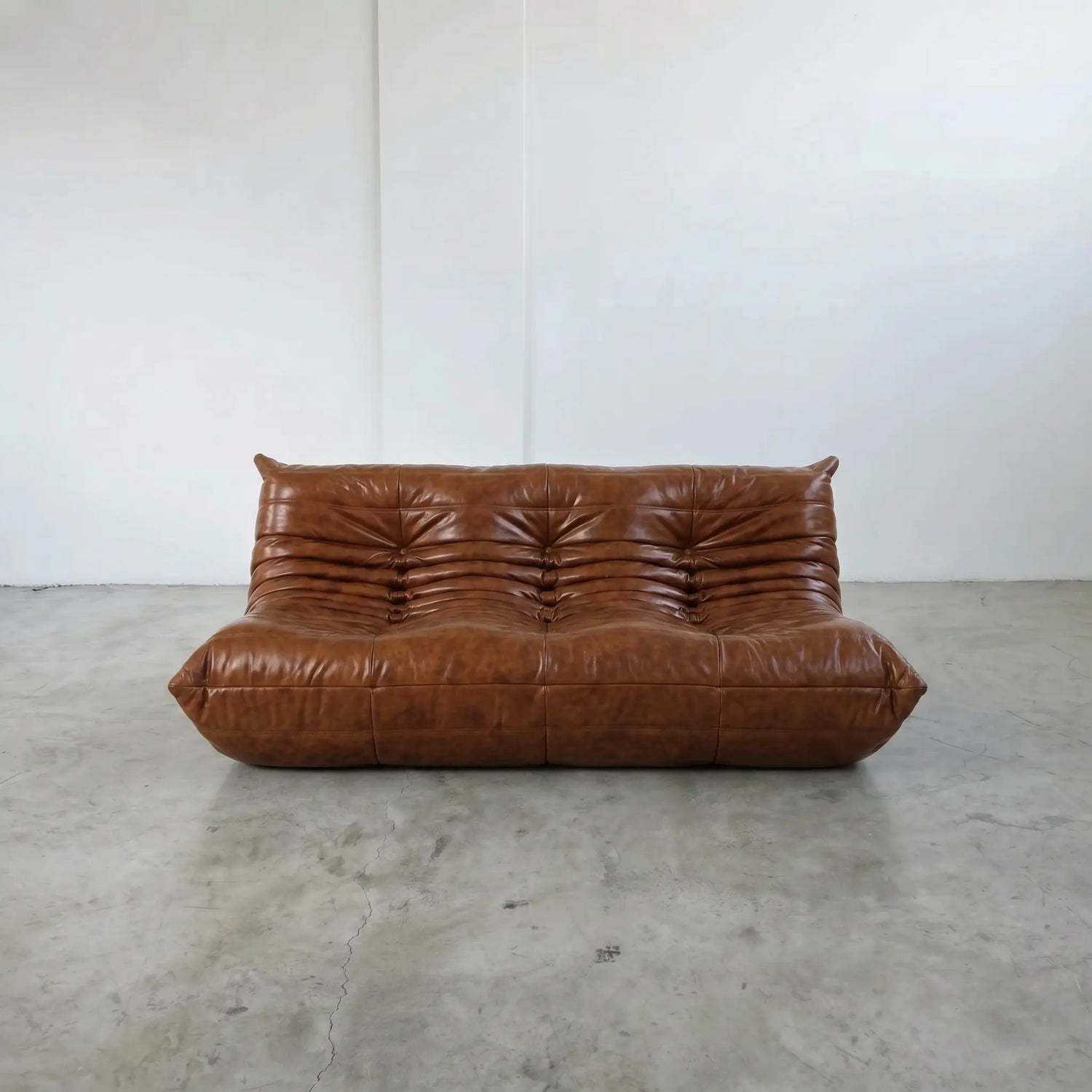 A togo sofa dupe in brown leather with a heavily tufted, channel-tufted backrest and thick cushioned arms, set against a minimalist white wall and concrete floor.