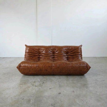 A togo sofa dupe in brown leather with a heavily tufted, channel-tufted backrest and thick cushioned arms, set against a minimalist white wall and concrete floor.