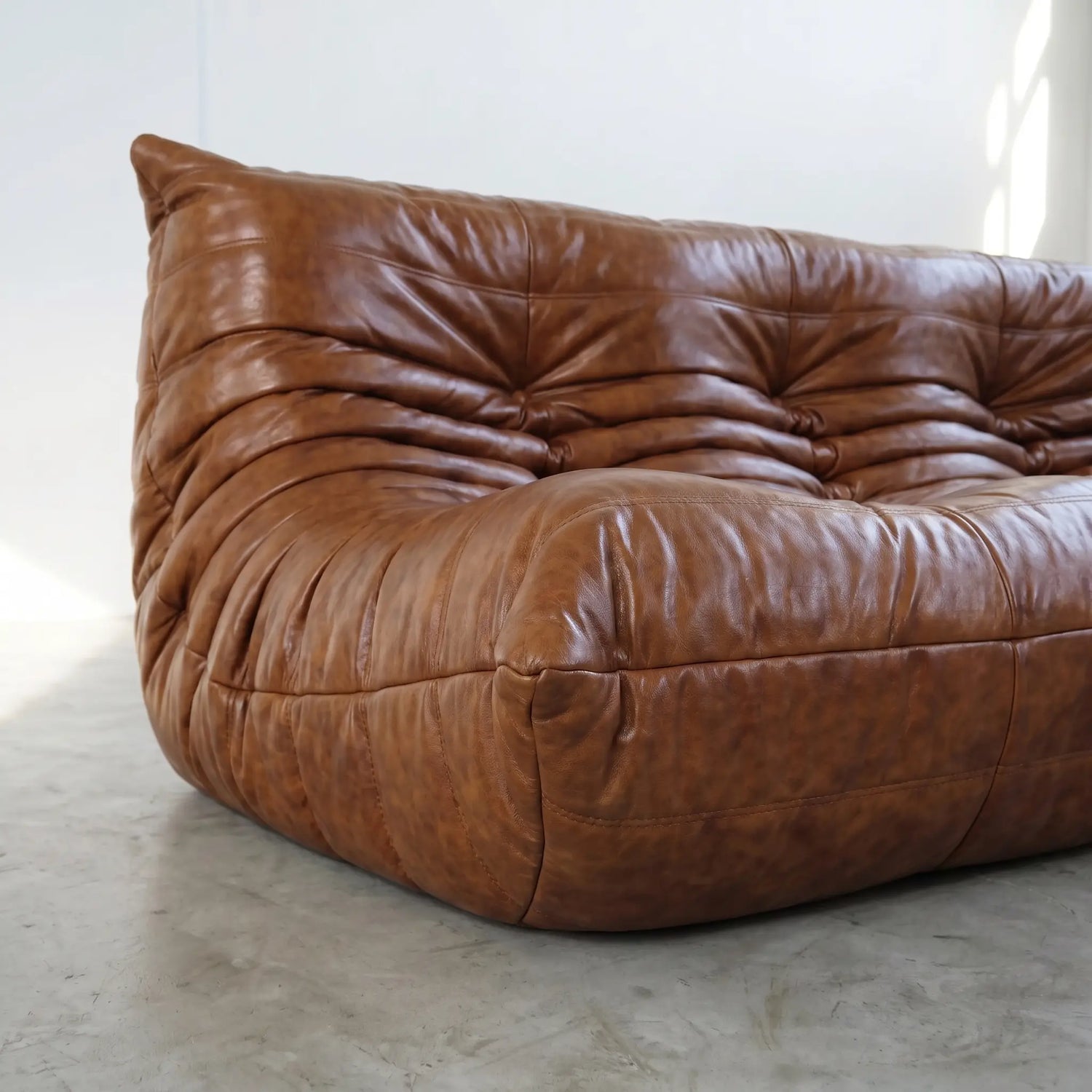 A close-up of a brown leather togo sofa dupe with a heavily tufted, cloud-like silhouette, set against a minimalist white wall and concrete floor.