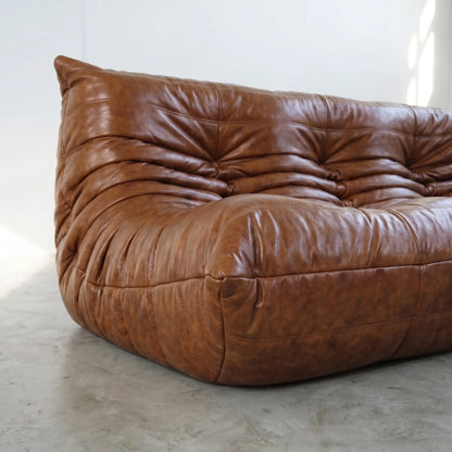 A close-up of a brown leather togo sofa dupe with a heavily tufted, cloud-like silhouette, set against a minimalist white wall and concrete floor.