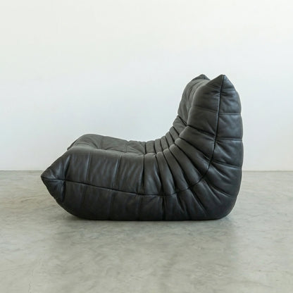 Modern togo chair replica in faux black leather on a gray floor with a white wall background