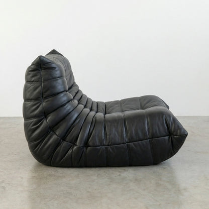 Togo Chair Dupe in black real cowhide leather on a concrete floor with a white wall background hot from a side angle