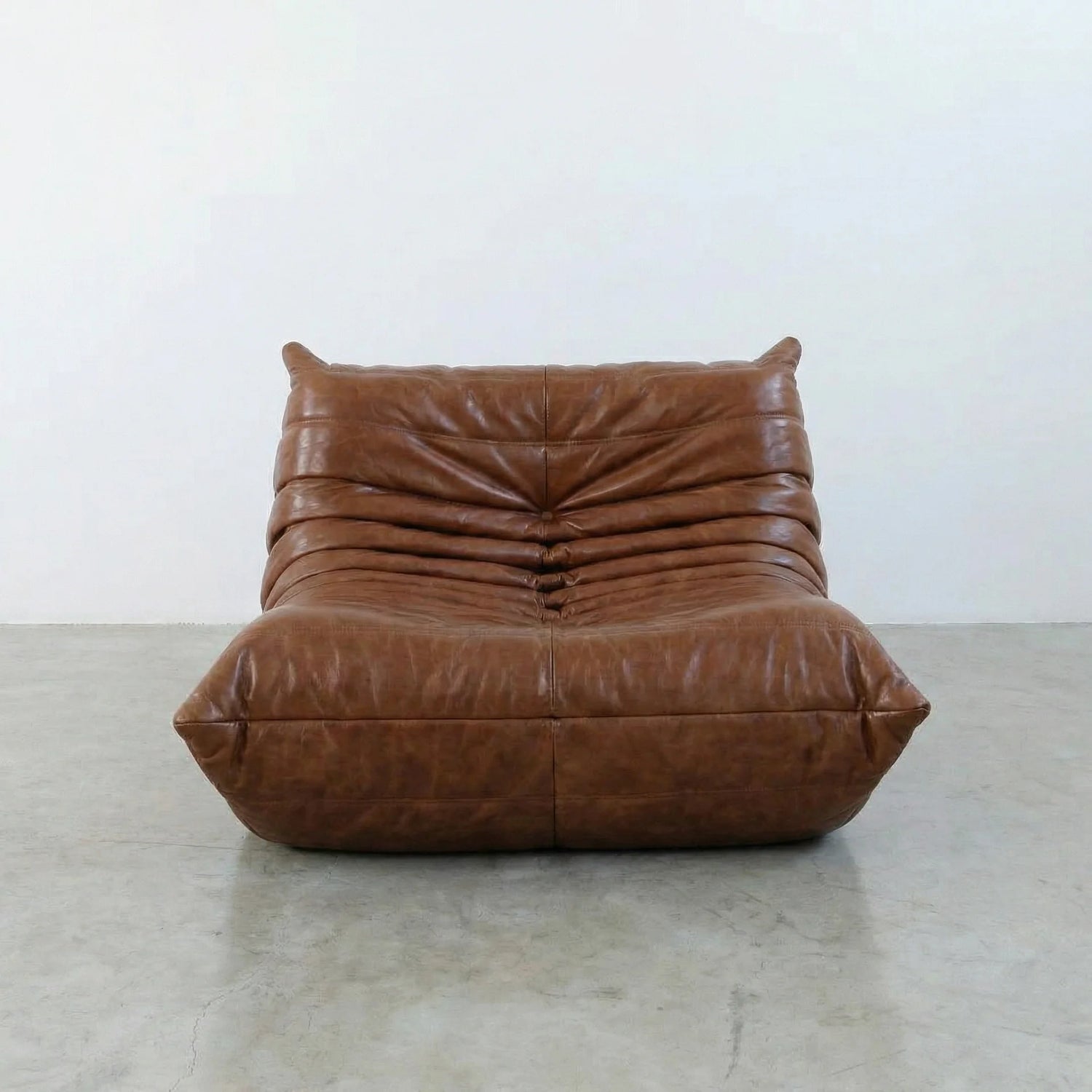 Togo Chair Dupe in brown real shiny oil wax leather on a concrete floor with a white wall background