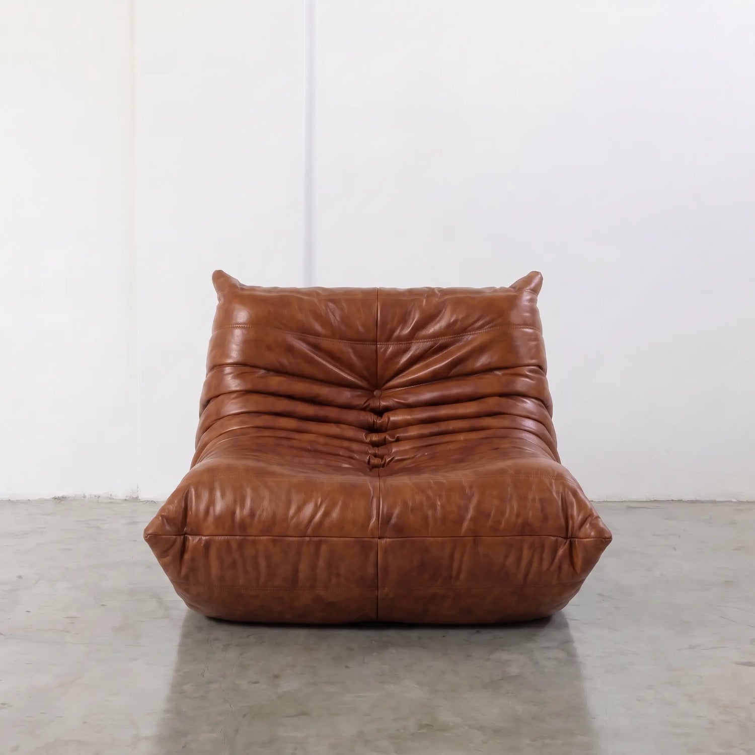 Togo Chair Dupe in brown real shiny oil wax leather on a concrete floor with a white wall background