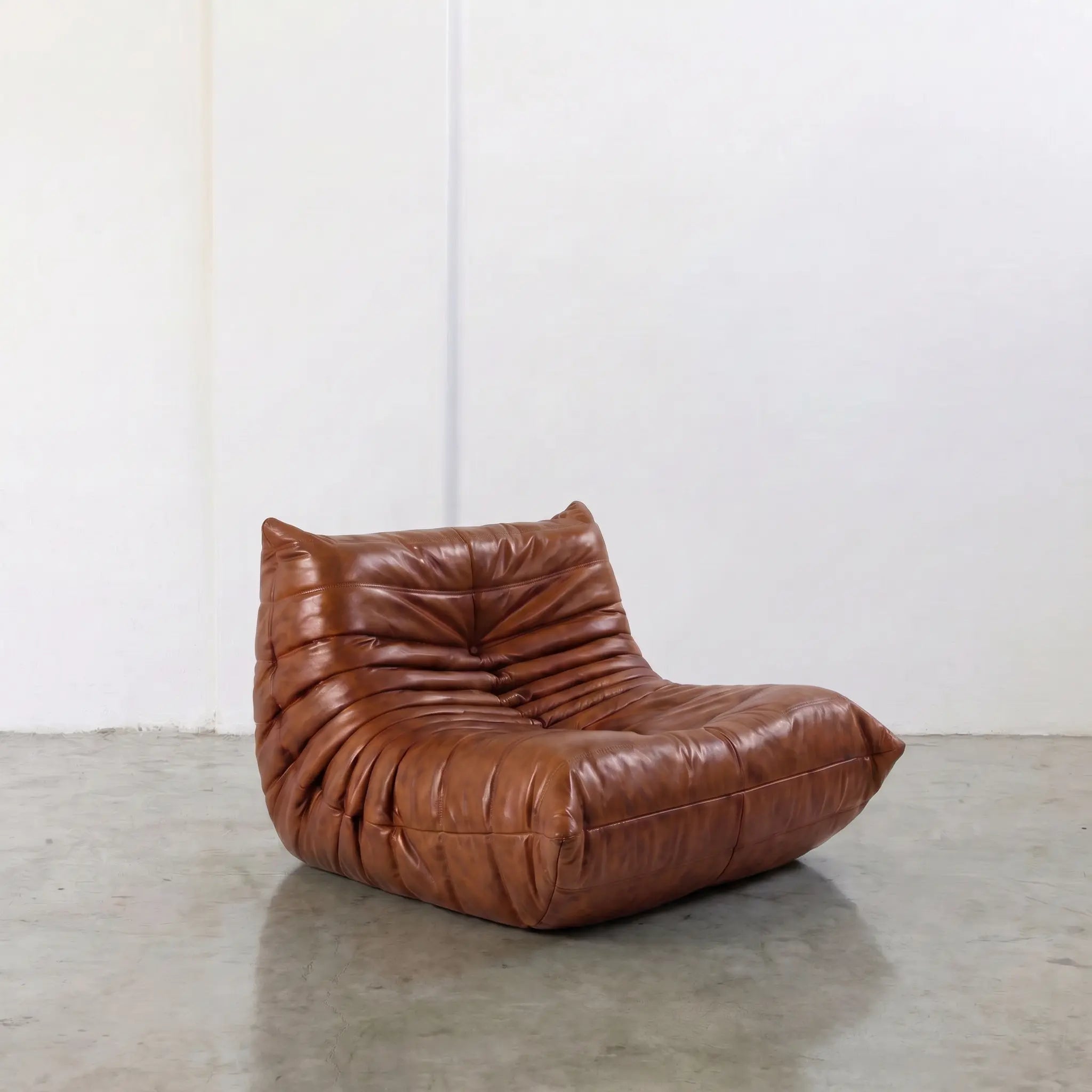 Togo Chair Dupe in brown real shiny oil wax leather on a concrete floor with a white wall background